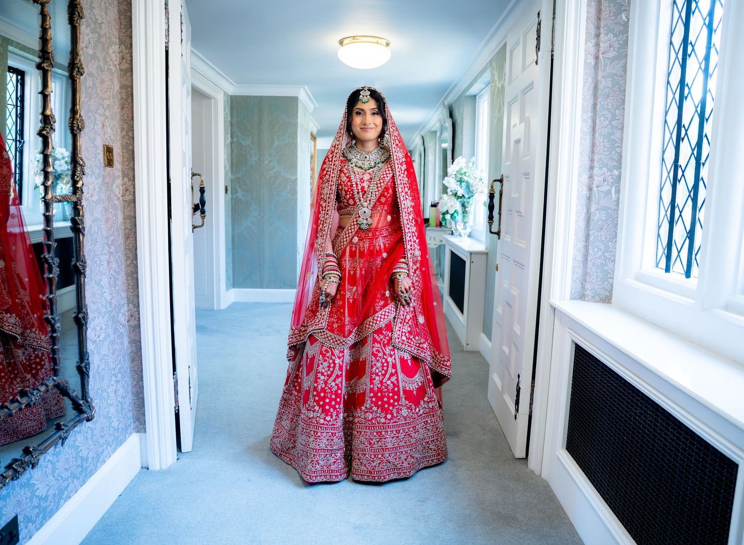 Indian bride in red embroidered lehenga with dupatta standing in bright hallway, full bridal look.