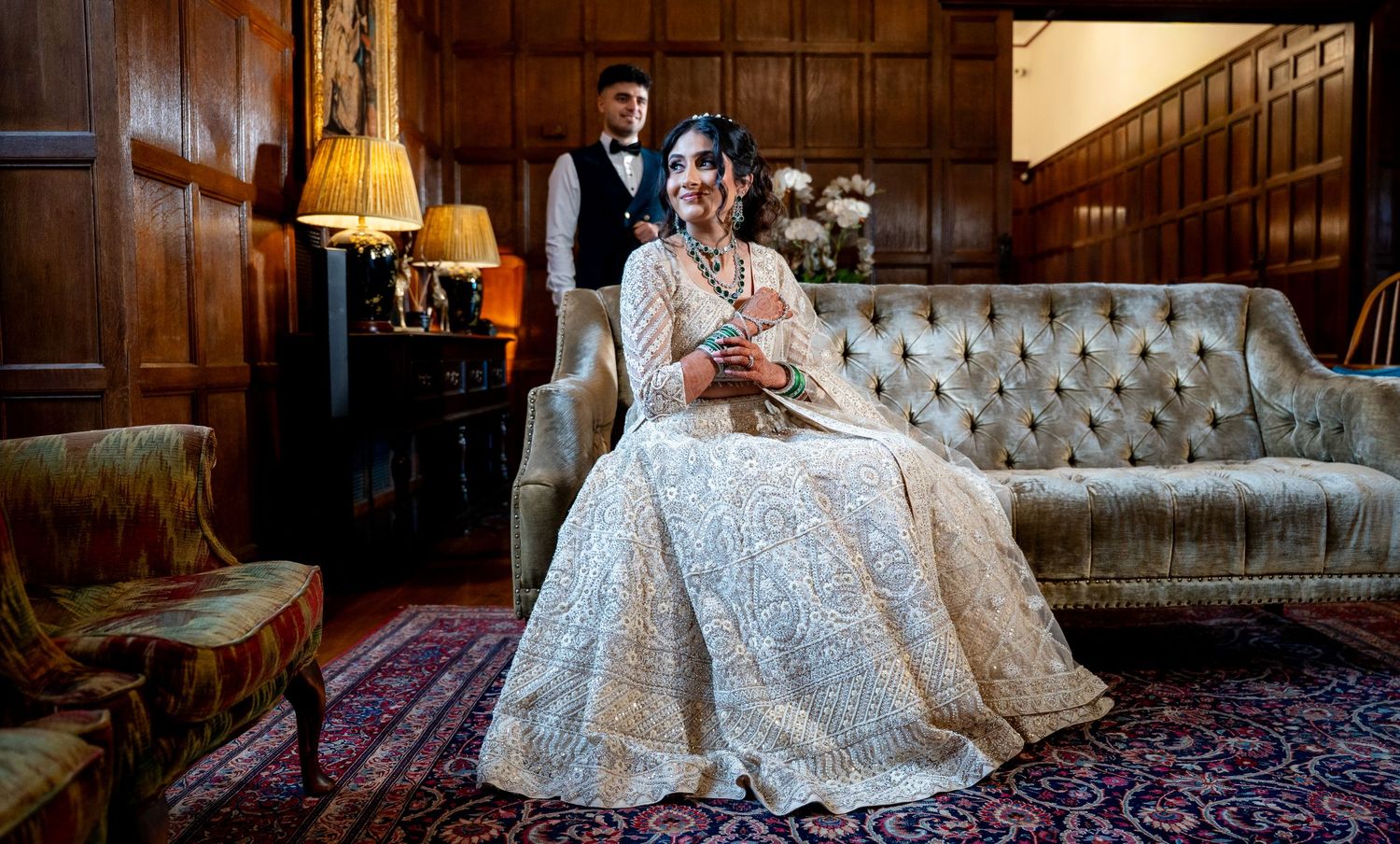 Bride in ornate white lehenga sits on vintage sofa as groom stands behind in elegant venue.