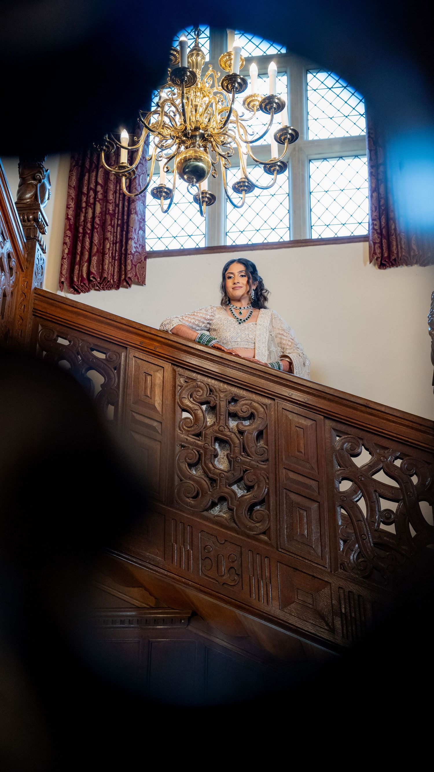 Bride in white lehenga stands at ornate wooden staircase balcony beneath golden chandelier in historic venue.