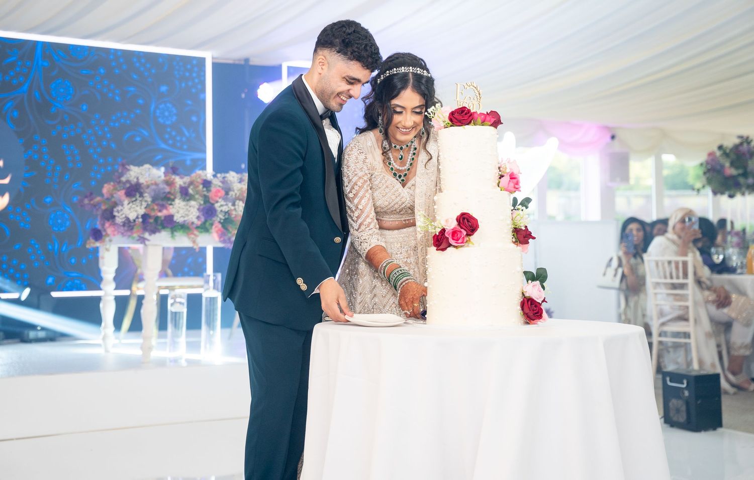 Smiling couple in South Asian wedding attire cutting elegant white rose-decorated wedding cake at reception.