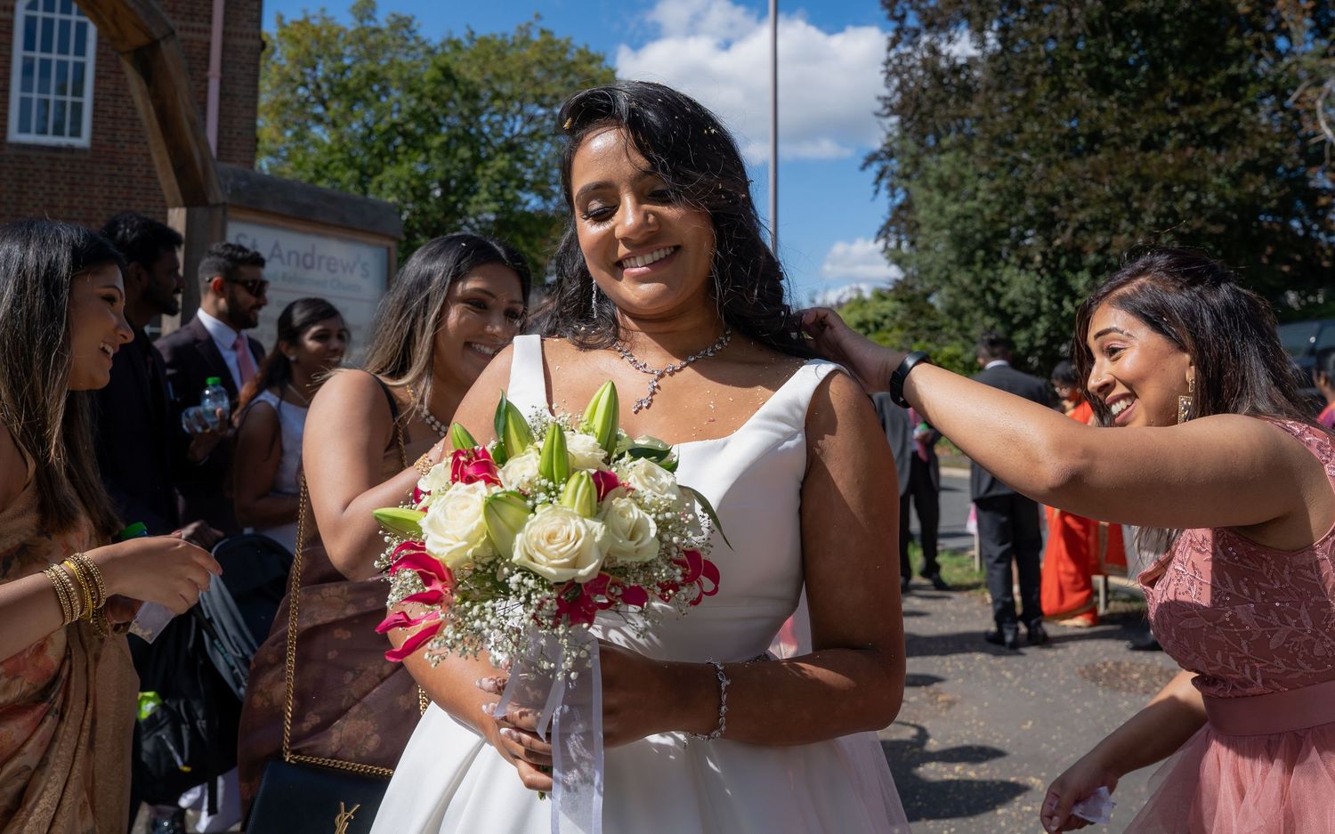 Bride in white dress holding bouquet of white and red flowers surrounded by smiling guests outdoors.
