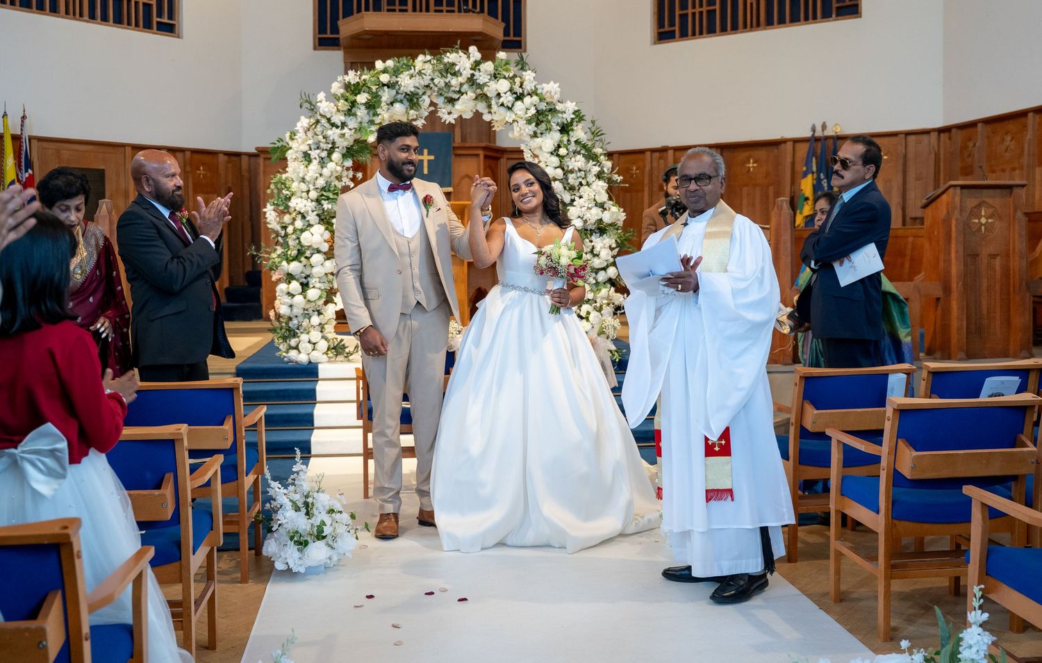 Newlyweds walk down church aisle after ceremony, officiant in white robes beside them, floral arch behind.