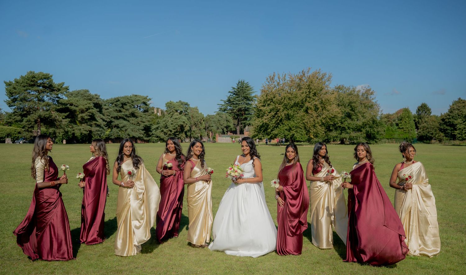 Bride in white gown poses with bridesmaids in gold and burgundy satin sarees on a lush green lawn.