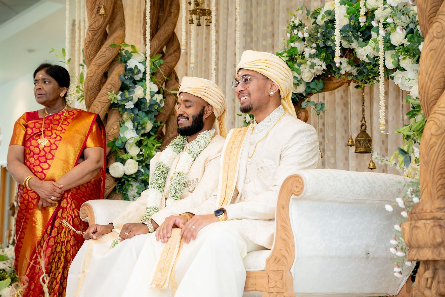 Two grooms in white sherwanis and turbans sit together at a South Asian wedding ceremony with floral decorations.