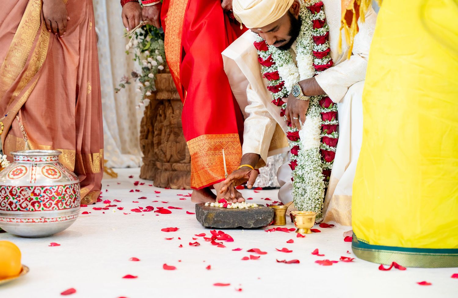 Close-up of South Indian wedding ritual with groom in white dhoti and bride in red saree performing sacred stone ceremony.