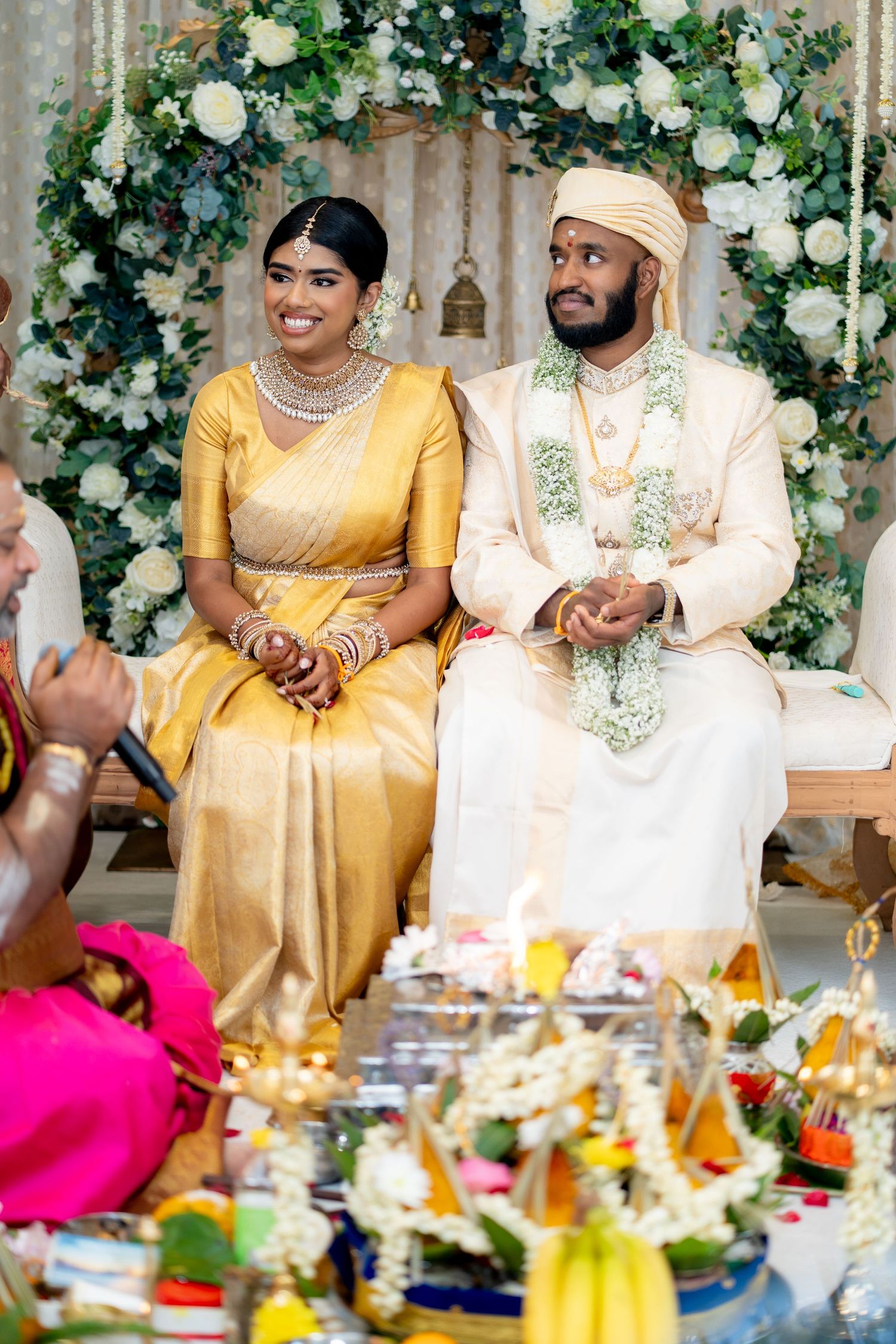 South Indian wedding couple in traditional attire seated at decorated mandap with ritual offerings arranged in foreground.
