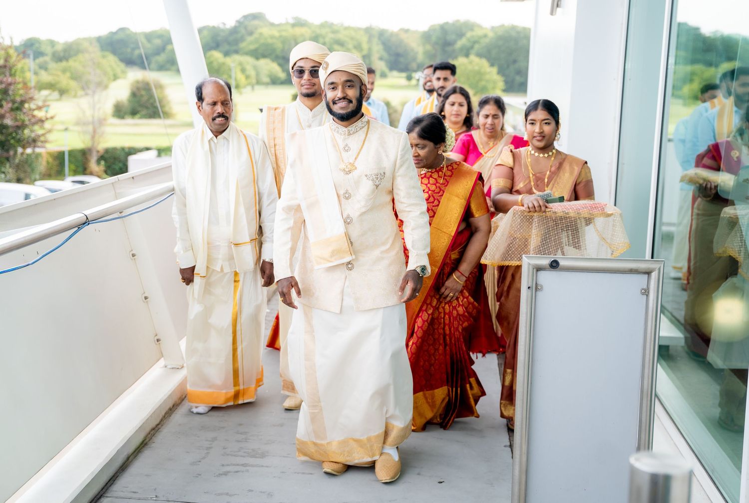 Indian wedding procession with grooms in white and gold traditional attire walking through a modern venue corridor.