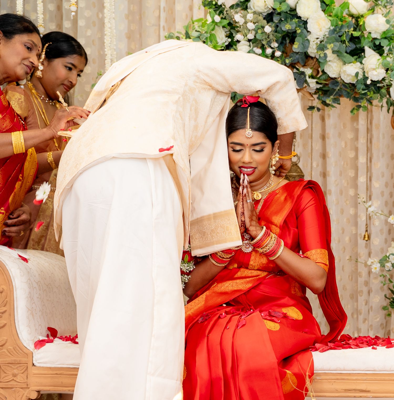 Groom in white dhoti blesses bride in red silk saree with hands in prayer during Hindu wedding ceremony.