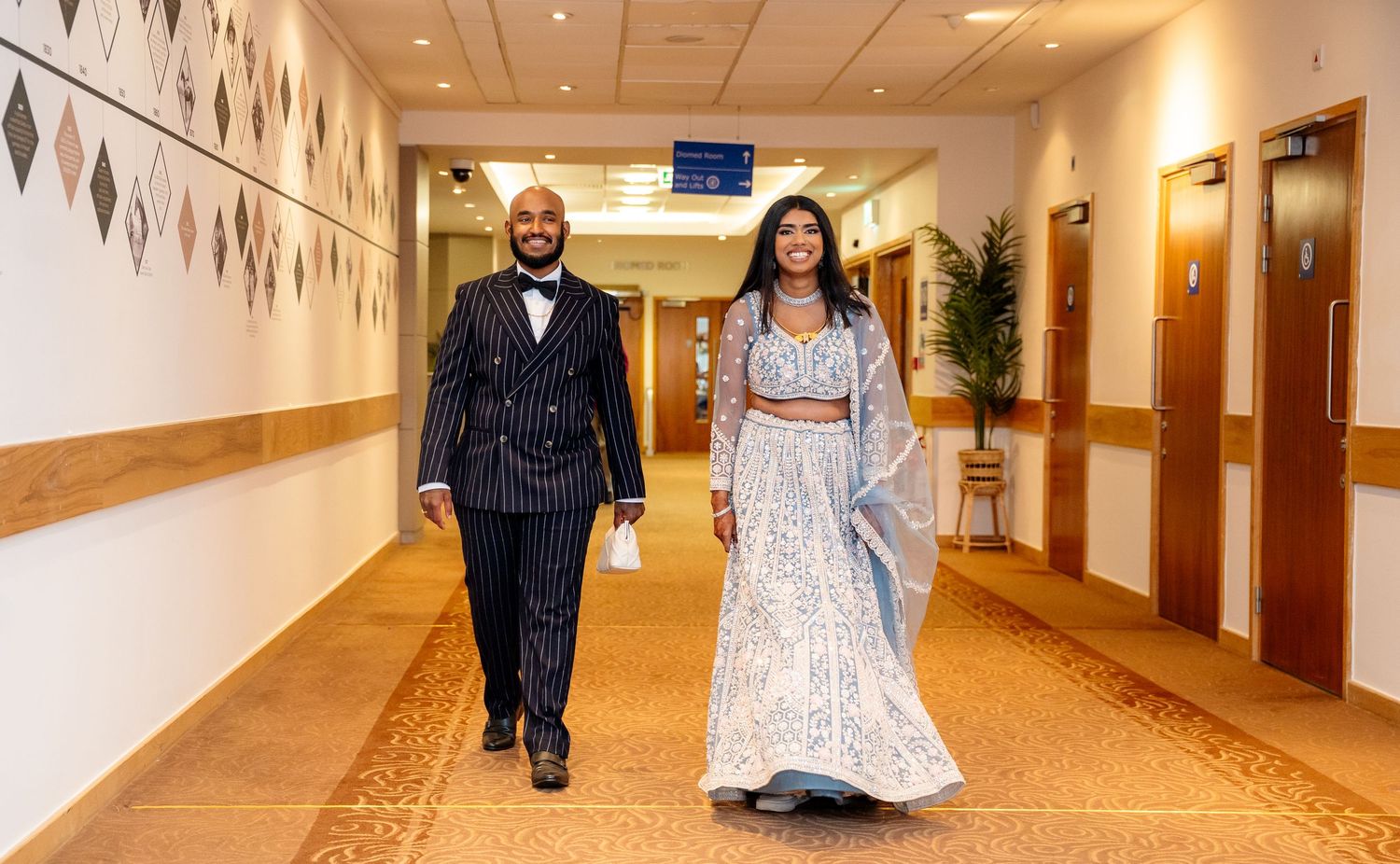 Elegant couple walking down hotel hallway, man in pinstripe suit and woman in blue embroidered lehenga.