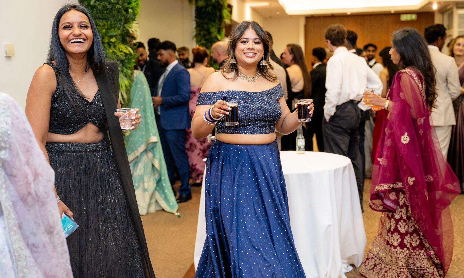 Two women smiling in Indian festive attire at a crowded wedding reception party.