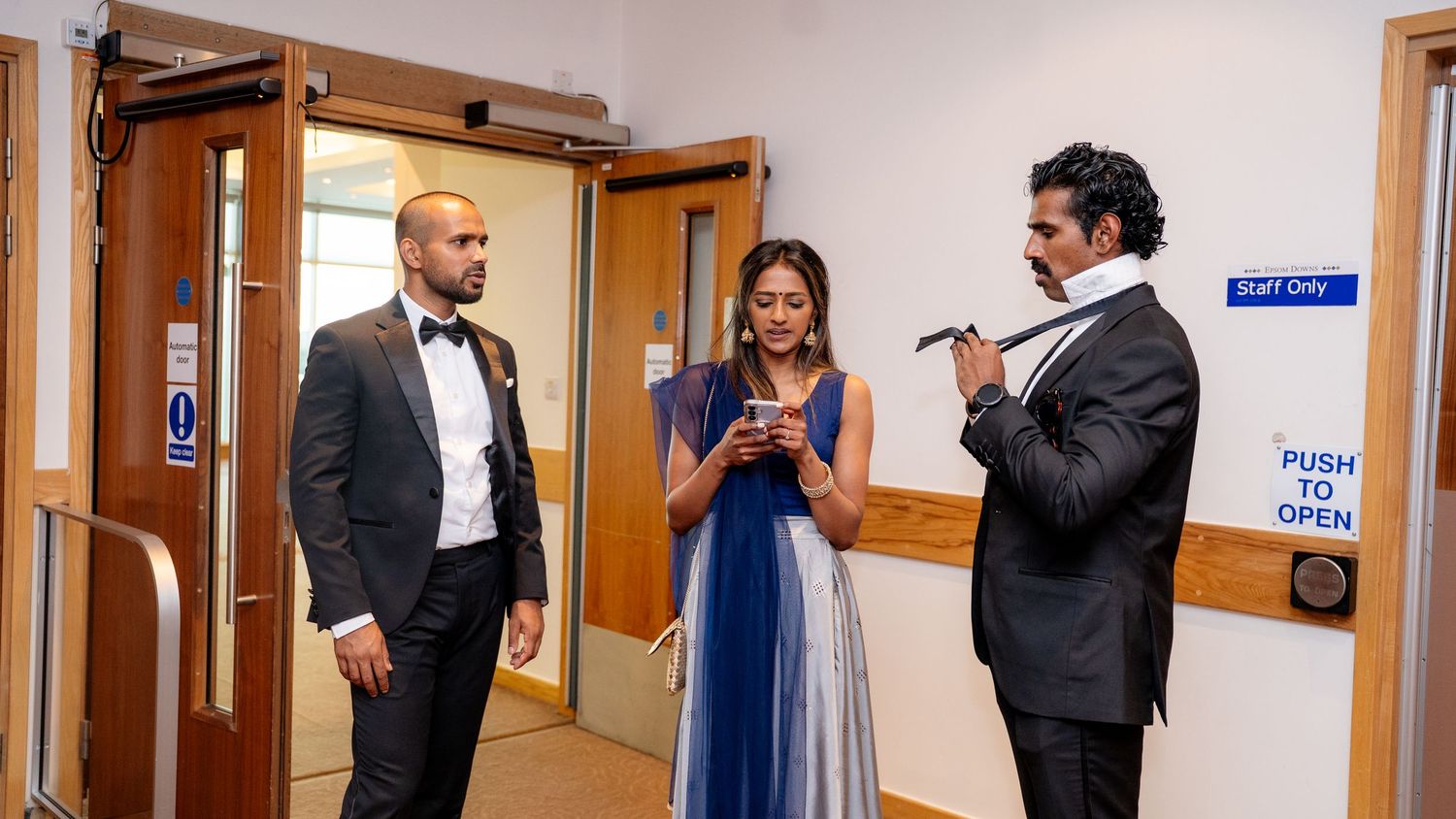 Three elegantly dressed guests conversing backstage at a formal event, with two men in tuxedos and a woman in a blue gown.