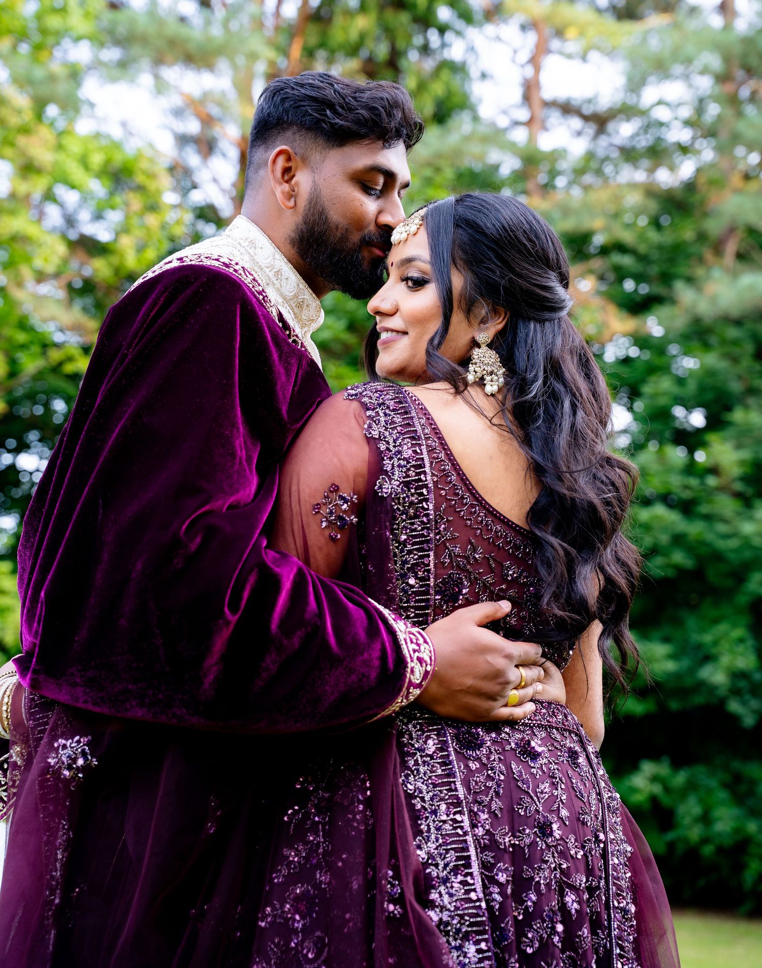 Groom in purple velvet sherwani kissing bride on forehead during romantic outdoor Indian wedding portrait.