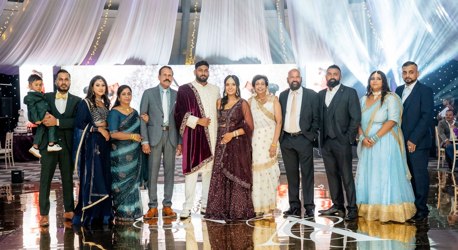 Large South Asian family group posing together at an elegant wedding reception in formal traditional attire.