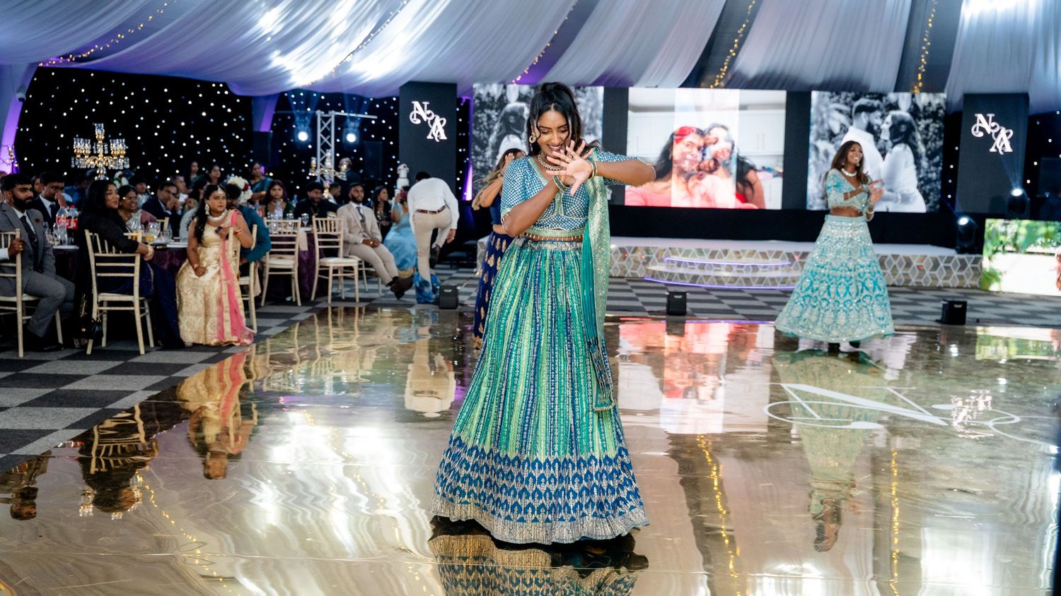 Woman in teal lehenga dancing at an Indian wedding reception with screens in background.