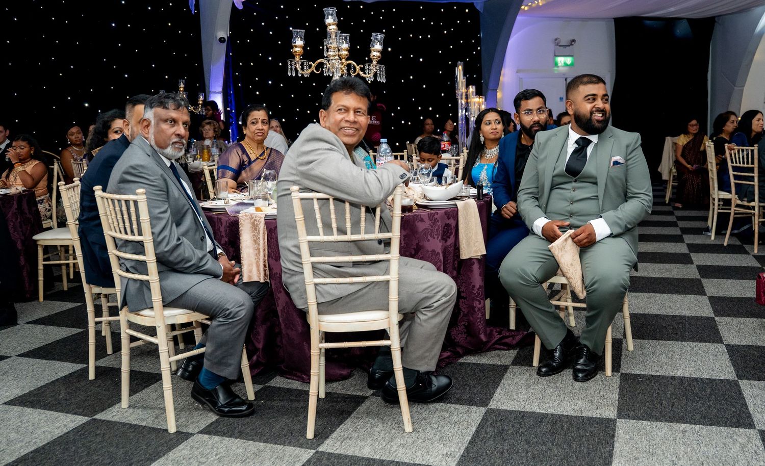 Men in suits seated at decorated wedding reception table with black and white checkered floor.