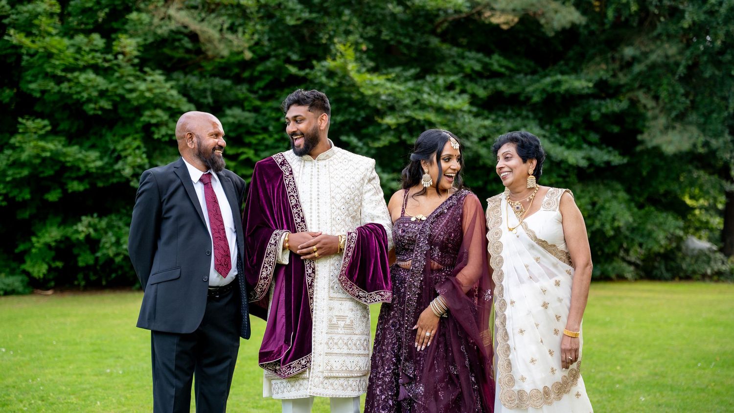 Indian wedding family of four laughing together outdoors, groom in white sherwani, bride in purple lehenga, on green lawn.