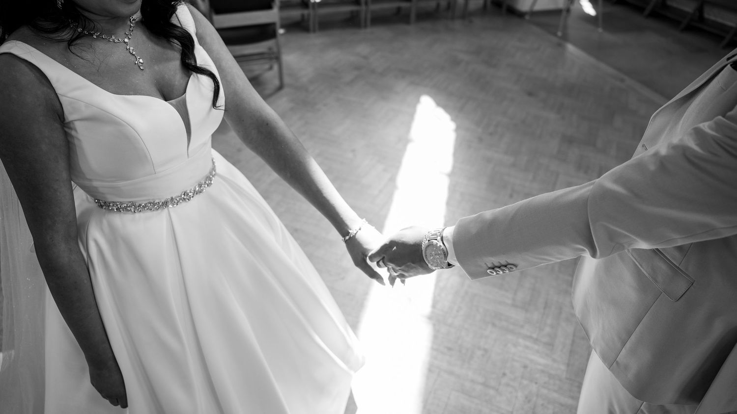 Bride and groom holding hands in elegant white wedding attire, captured in black and white photography.