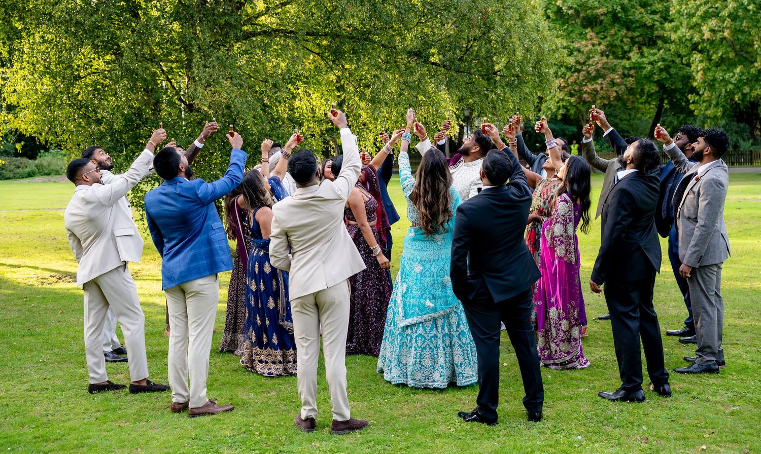 Wedding guests in colorful Indian and formal attire raising hands together in celebration on a lush green lawn.