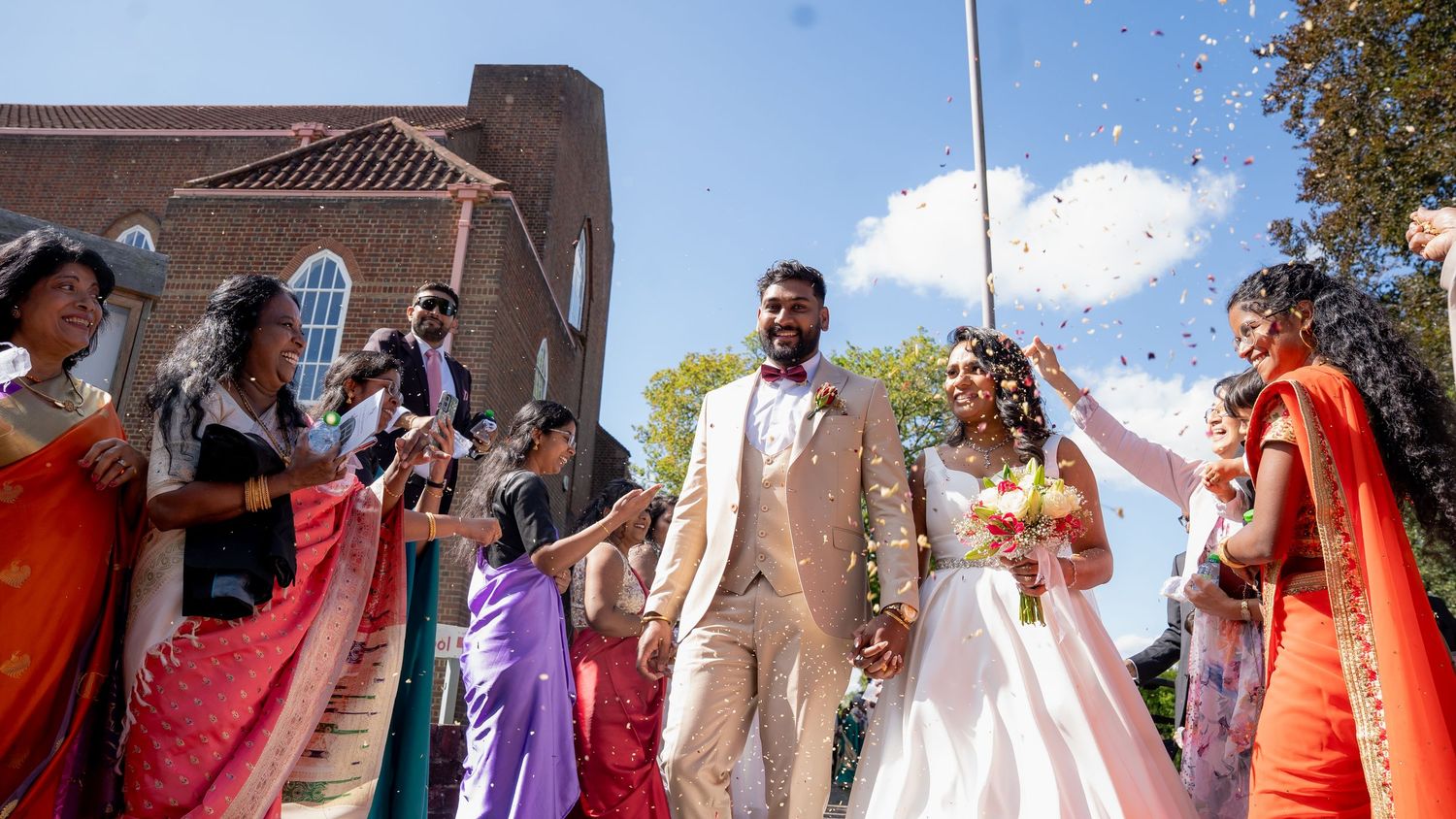 Indian wedding couple walks through confetti shower outdoors, surrounded by guests in colorful saris near a brick church.