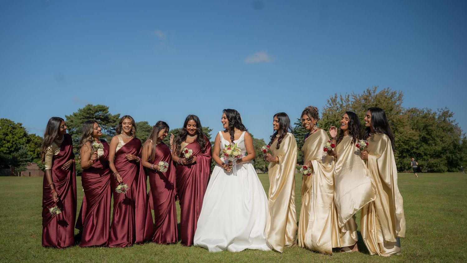 Bride in white gown laughing with bridesmaids in burgundy and gold sarees on sunny lawn.