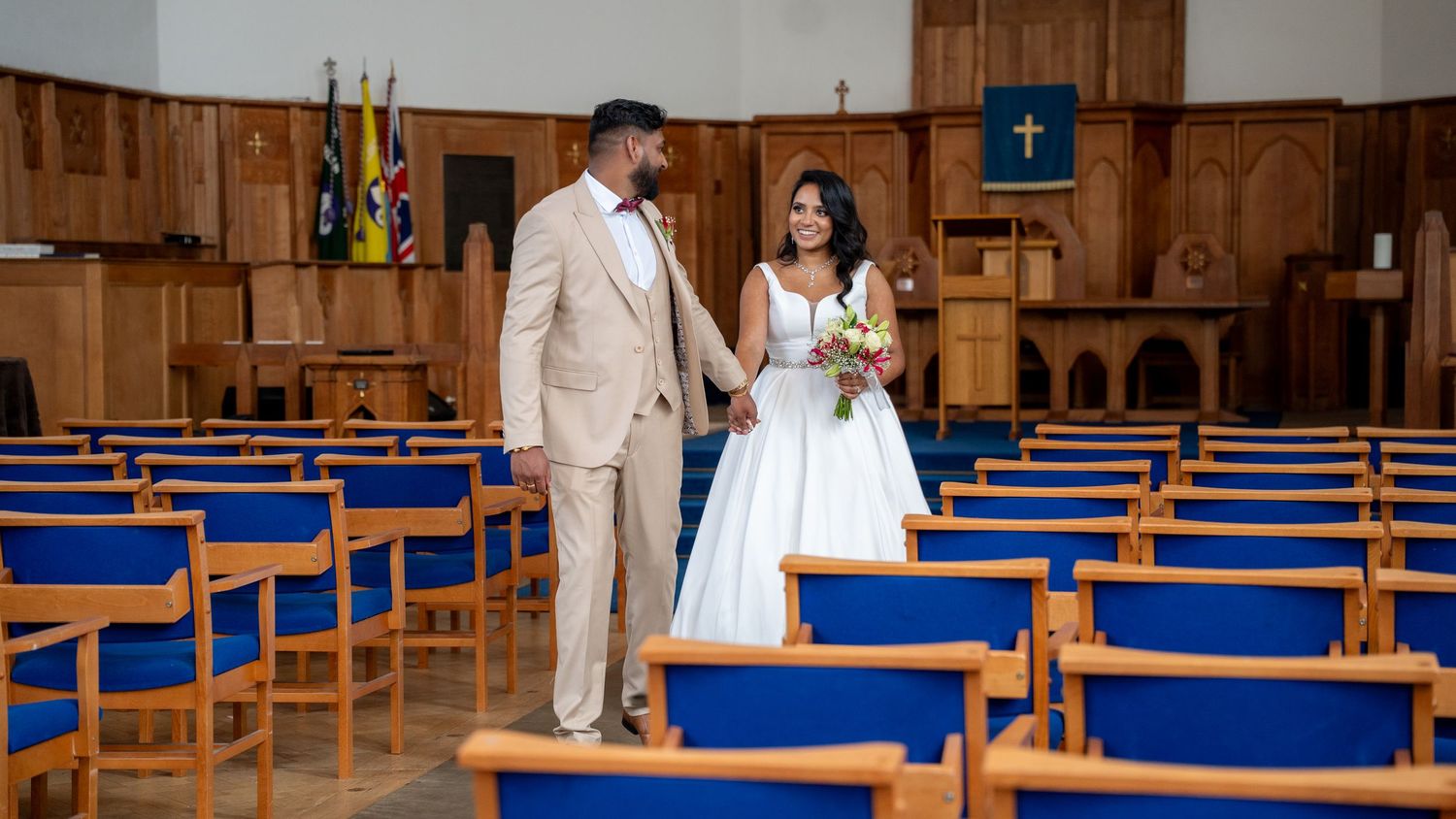 Newlywed couple walks hand-in-hand down church aisle, groom in beige suit, bride in elegant white wedding gown.