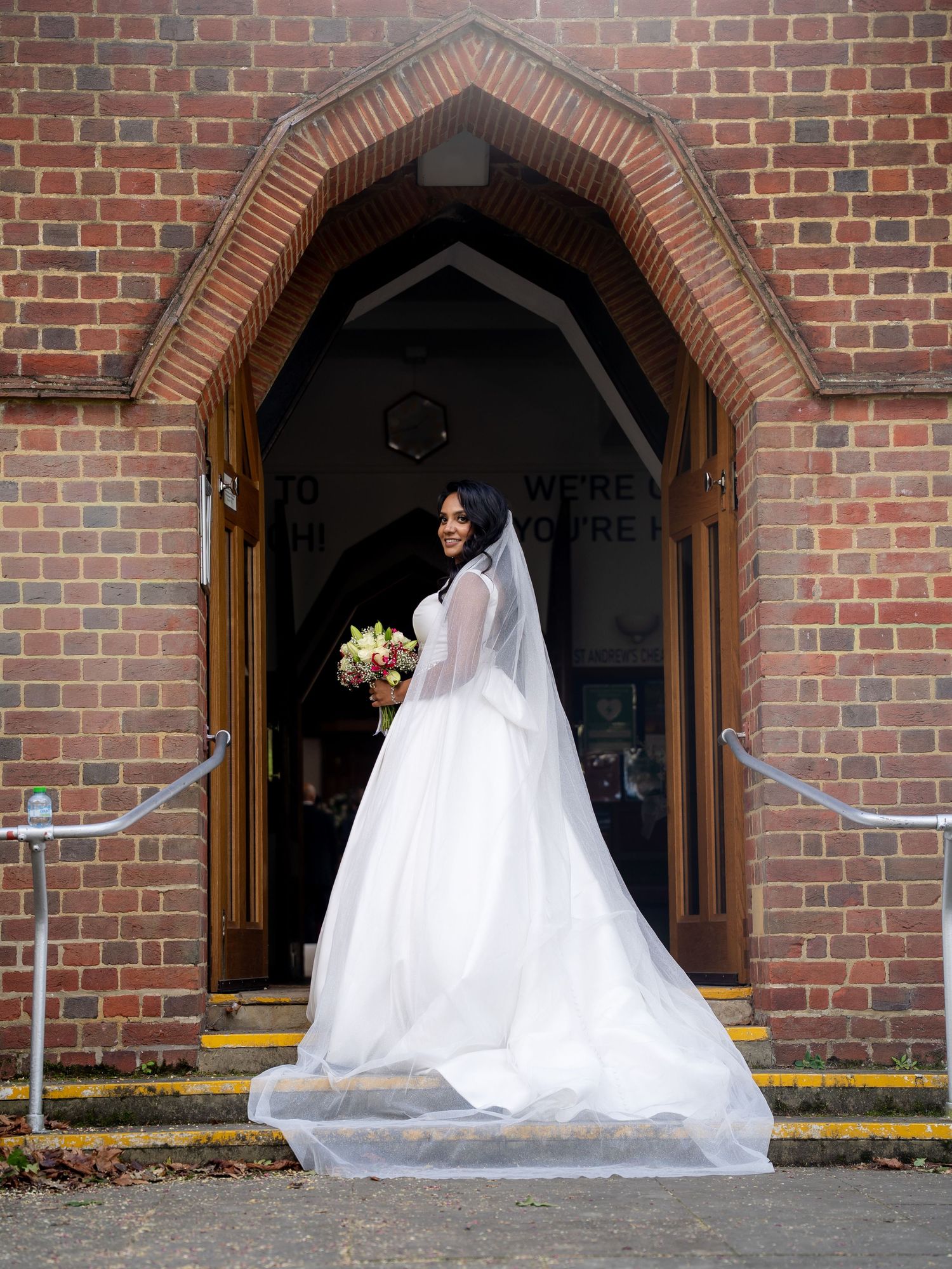 Bride in white ball gown with long veil holding bouquet at brick church entrance on wedding day.