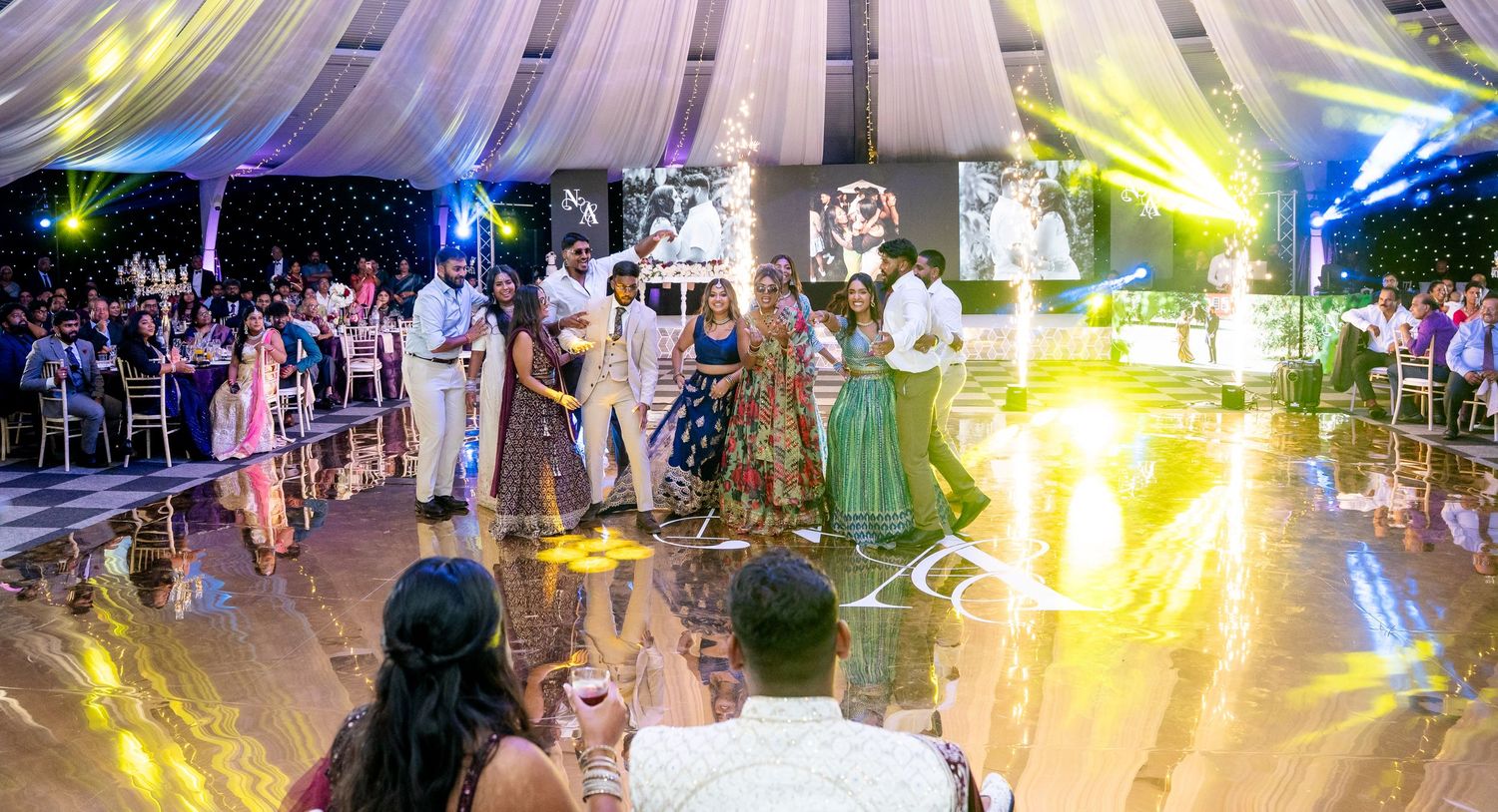 Guests dancing and celebrating at an elegant wedding reception with colorful lights and draped ceiling decor.