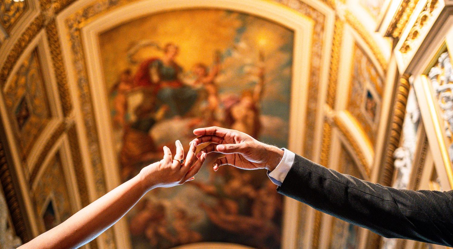 Close-up of hands exchanging wedding ring under ornate golden painted church ceiling.