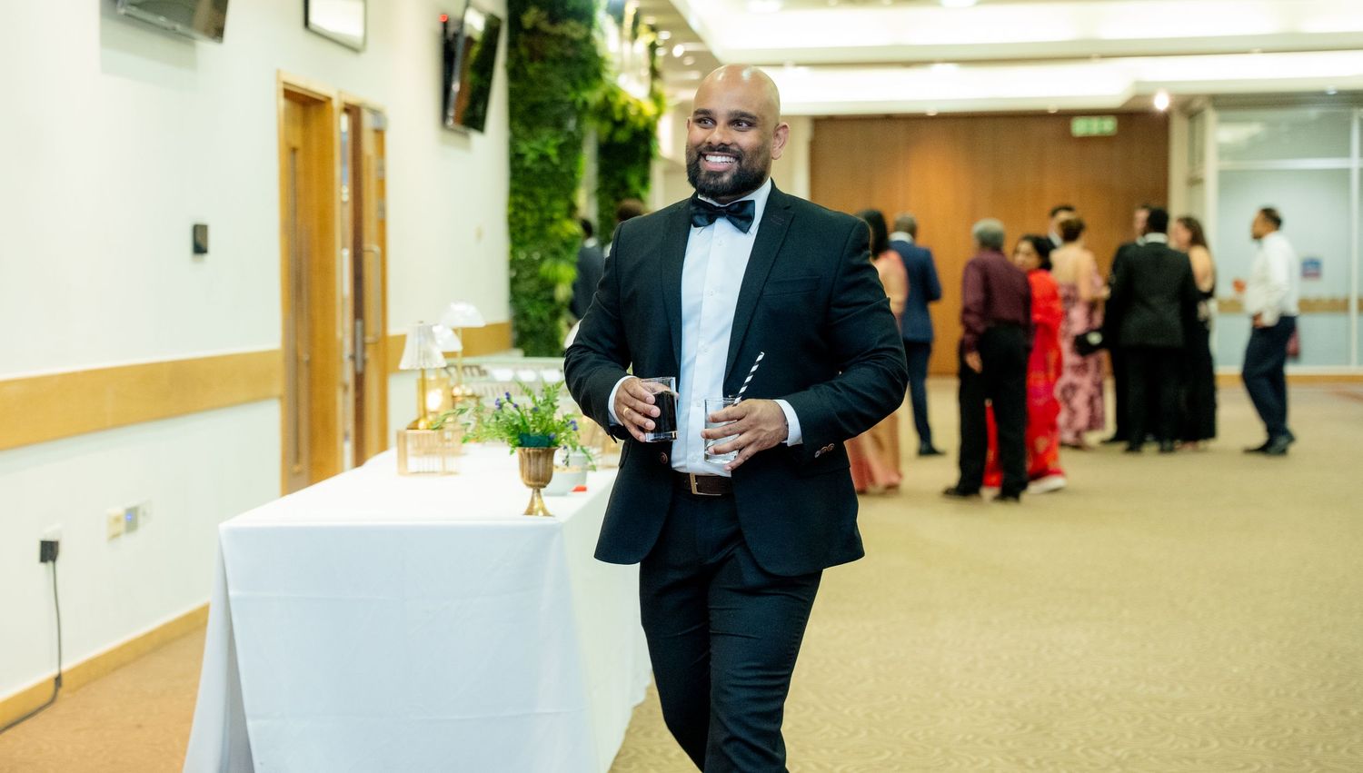 Smiling man in black tuxedo with bow tie walking through a formal event venue.