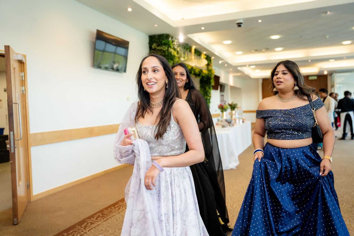 Women in elegant Indian traditional lehenga outfits walking through a bright wedding reception hall.