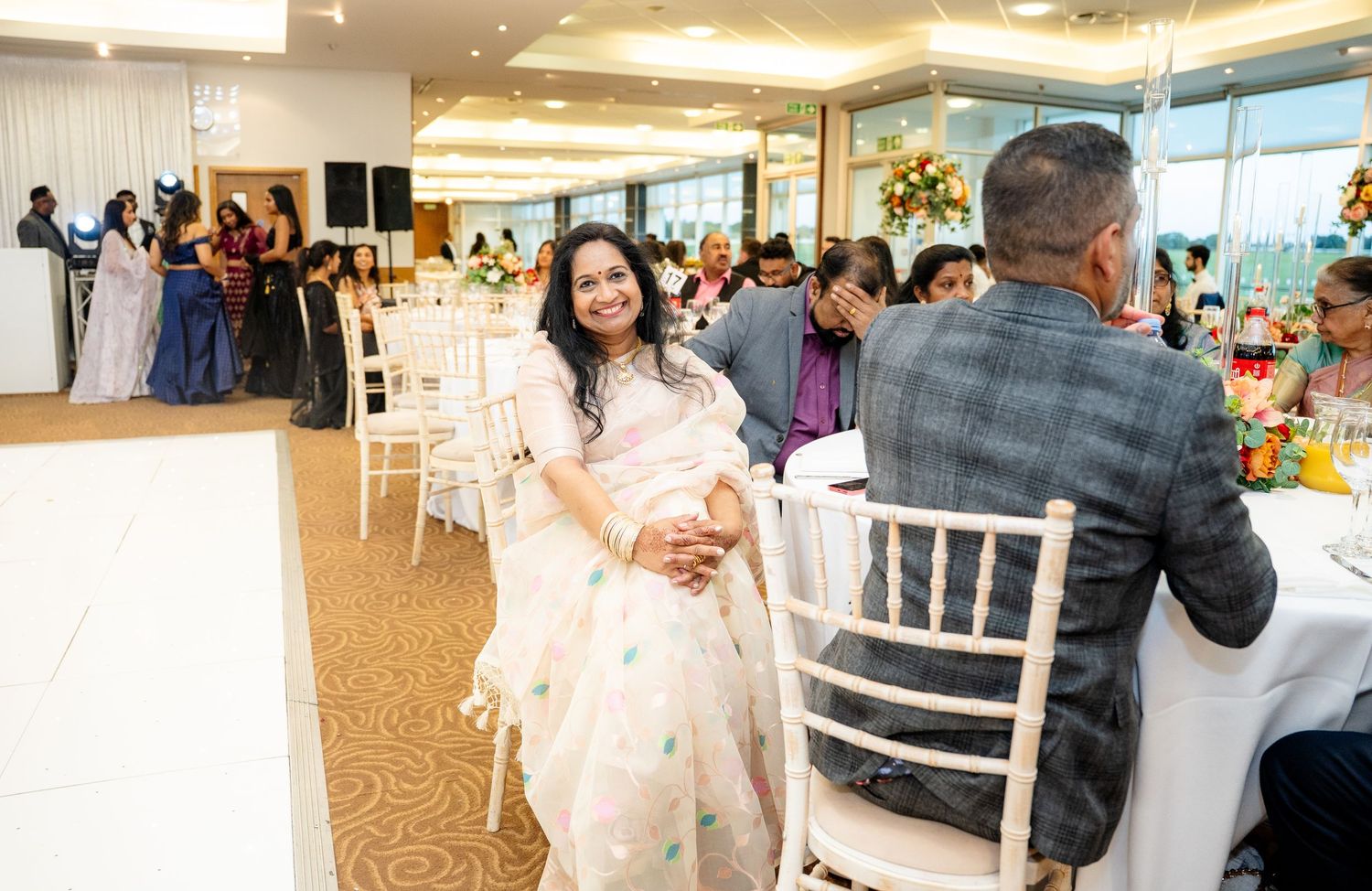 Woman in white saree smiles while seated at a beautifully decorated wedding reception hall.