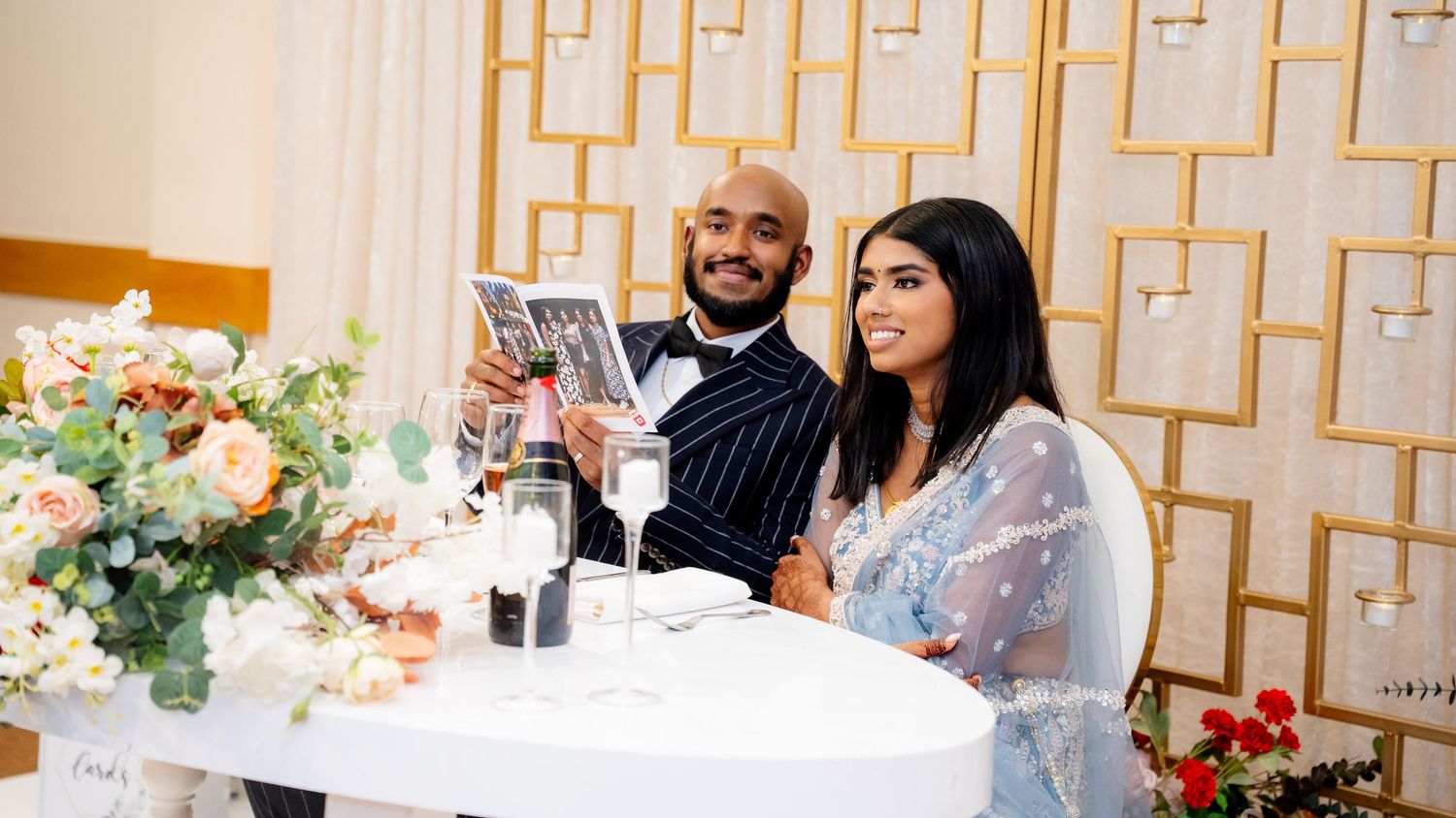 A smiling couple in wedding attire poses at a floral-decorated sweetheart table holding a photo.