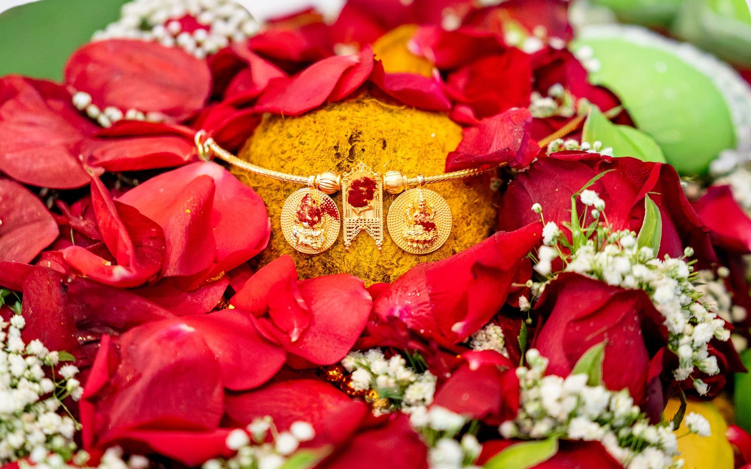Gold bangle with red gemstones displayed on turmeric surrounded by red rose petals and white baby's breath flowers.