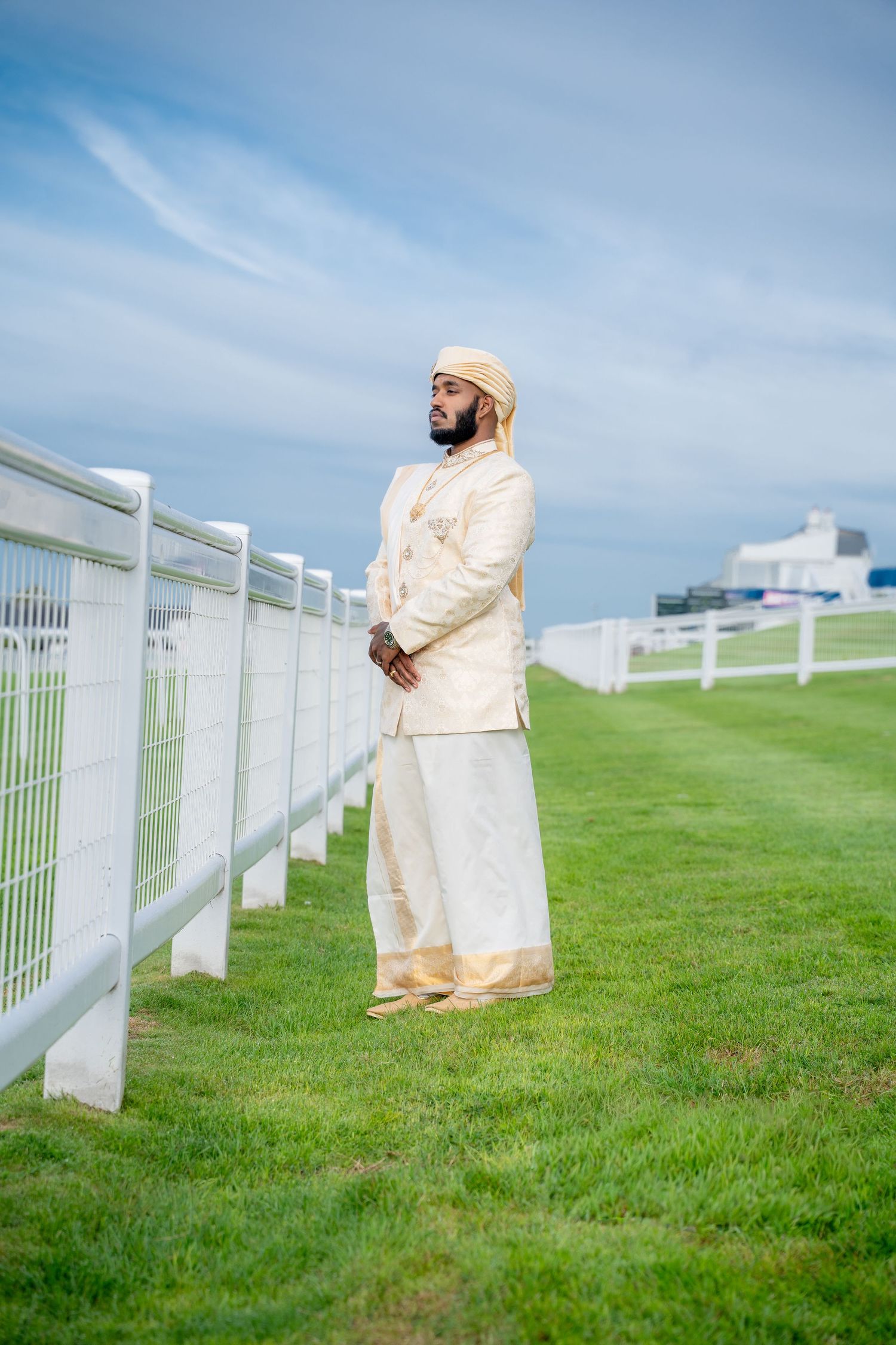 Indian groom in ivory gold sherwani and turban standing beside white fence at a racecourse.