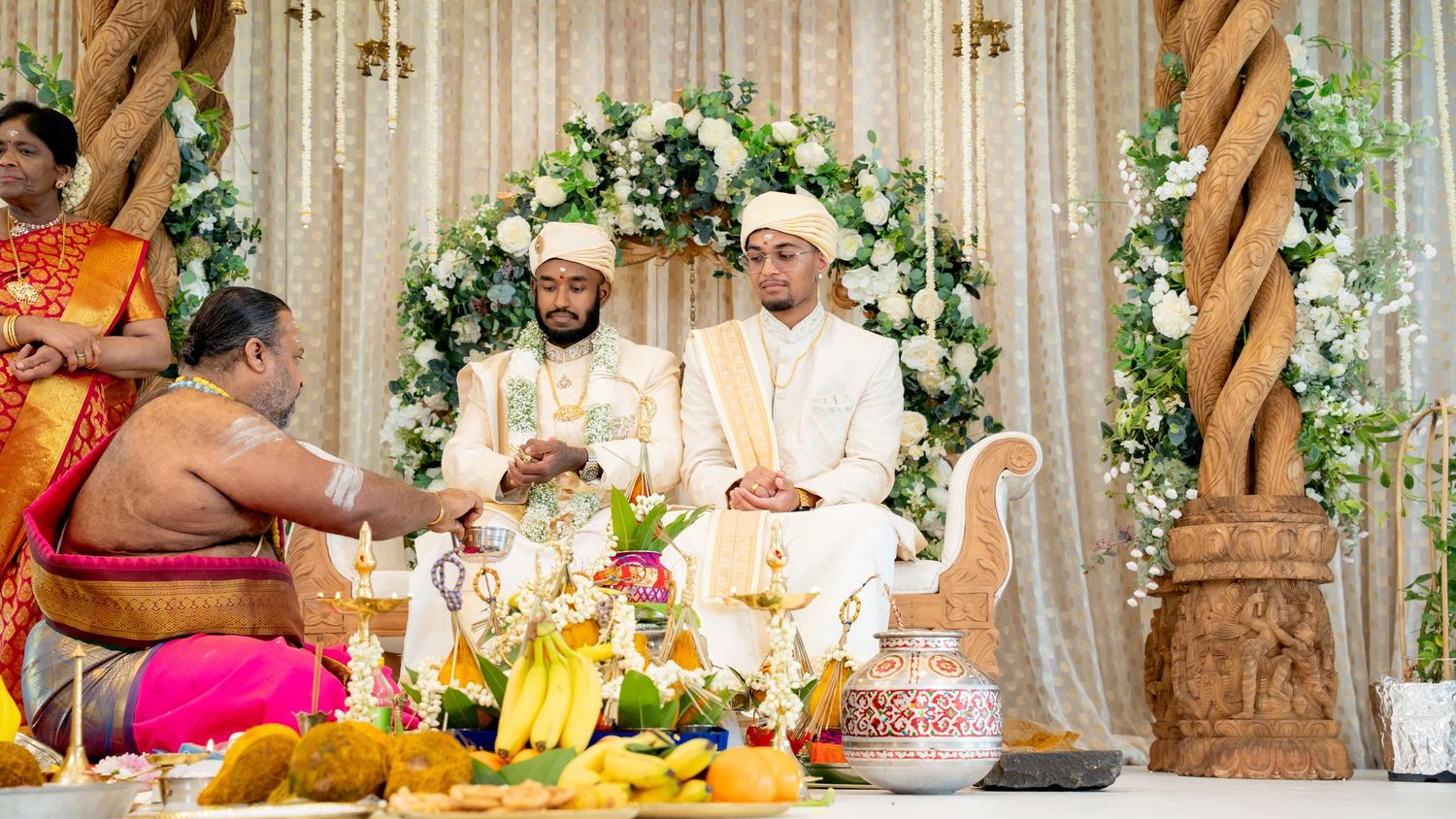 Two grooms in white attire participate in a traditional Hindu wedding ceremony with a priest.