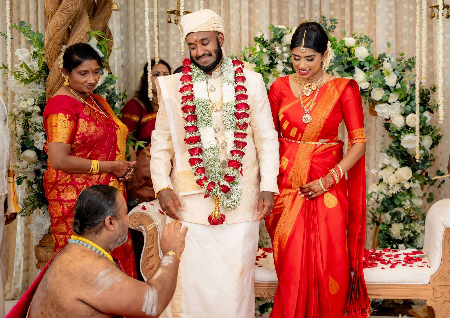 Groom in cream sherwani and bride in red silk saree stand together during South Indian wedding ritual.