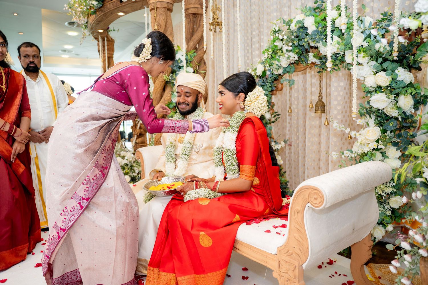 Family member blessing smiling Hindu bride and groom seated at elegant floral decorated wedding mandap.