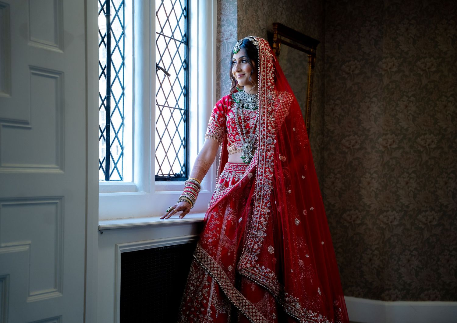 Indian bride in red bridal lehenga gazing out latticed window in a classic interior setting.