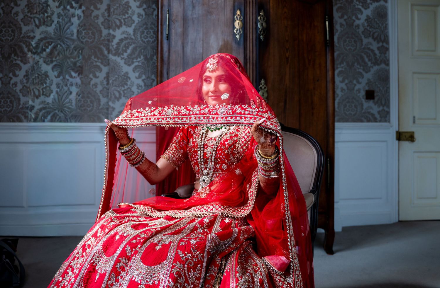 Smiling Indian bride in red lehenga holding dupatta veil in ornate room with traditional jewelry.