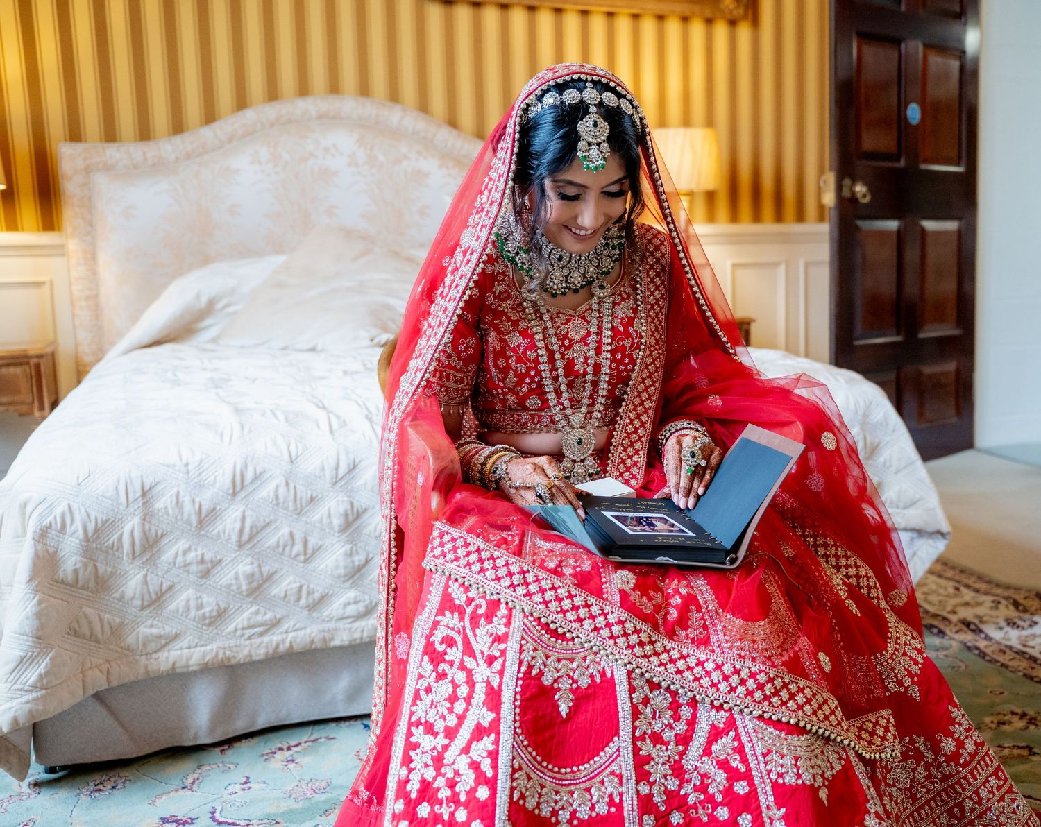 Indian bride in red lehenga with gold embroidery sits on bed using laptop on her wedding day.