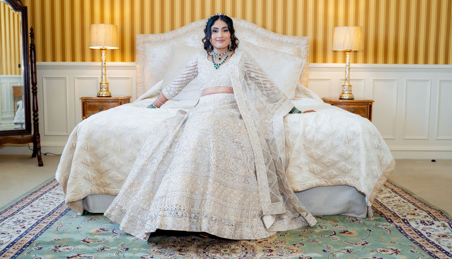 Indian bride in ivory embroidered lehenga seated on bed in luxurious hotel room.