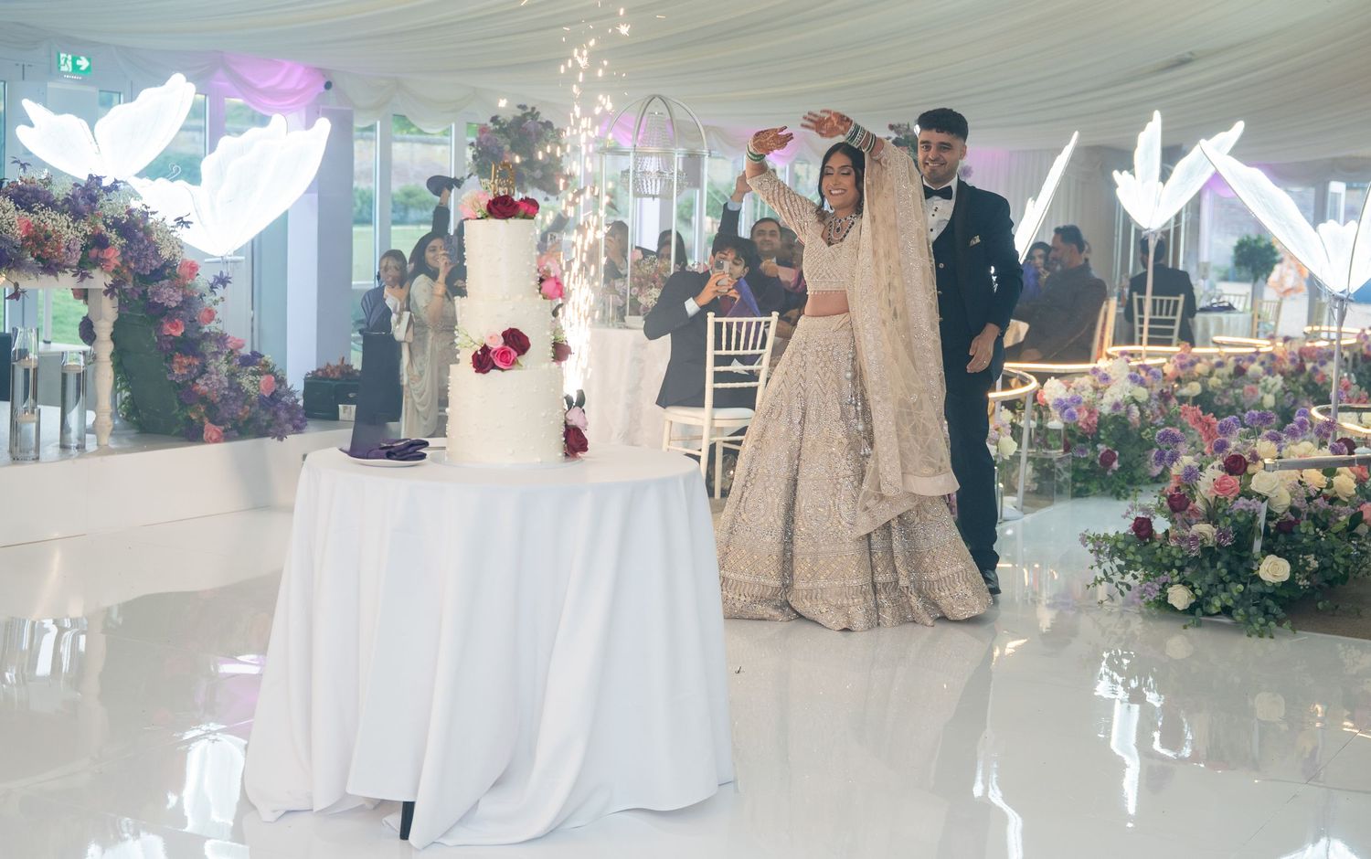 Bride and groom in traditional South Asian attire cutting a white wedding cake with sparklers at an elegant floral reception.