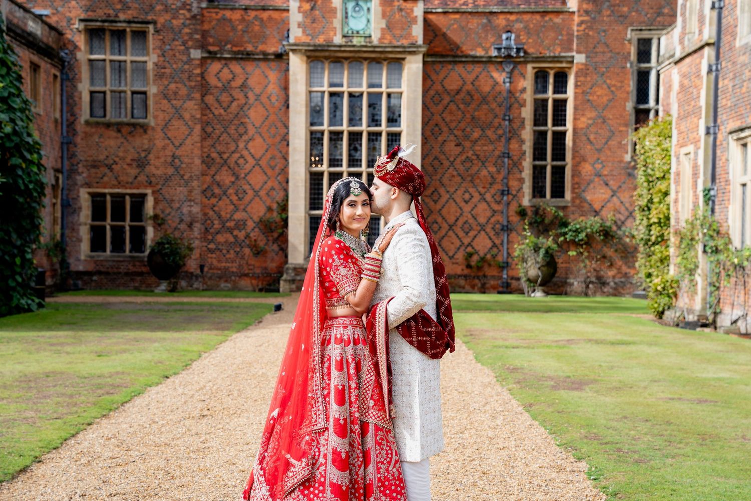 Indian wedding couple in red bridal lehenga and white sherwani sharing a tender moment at historic manor.