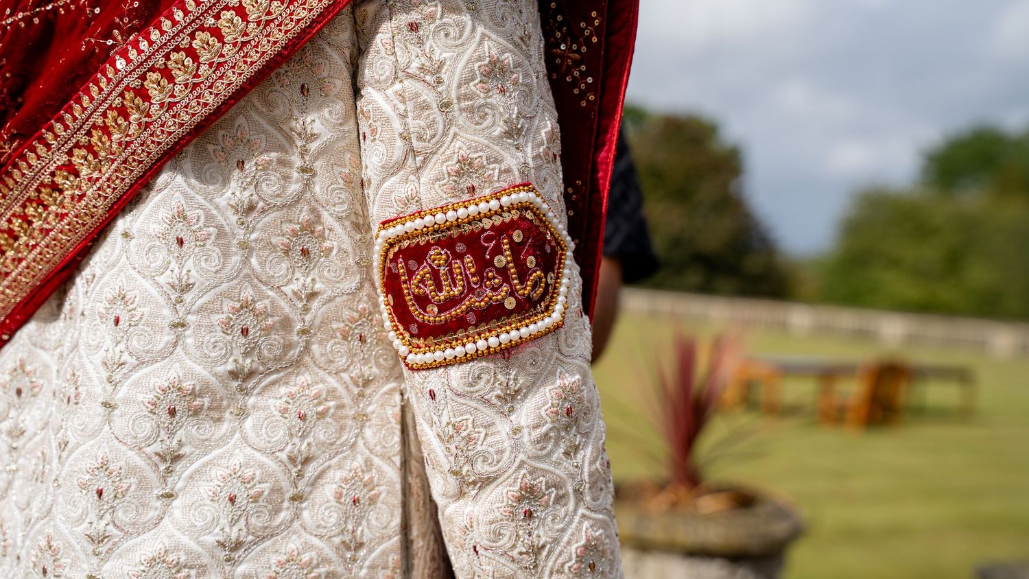 Close-up of intricate white embroidered bridal outfit with red and gold Arabic calligraphy patch detail.