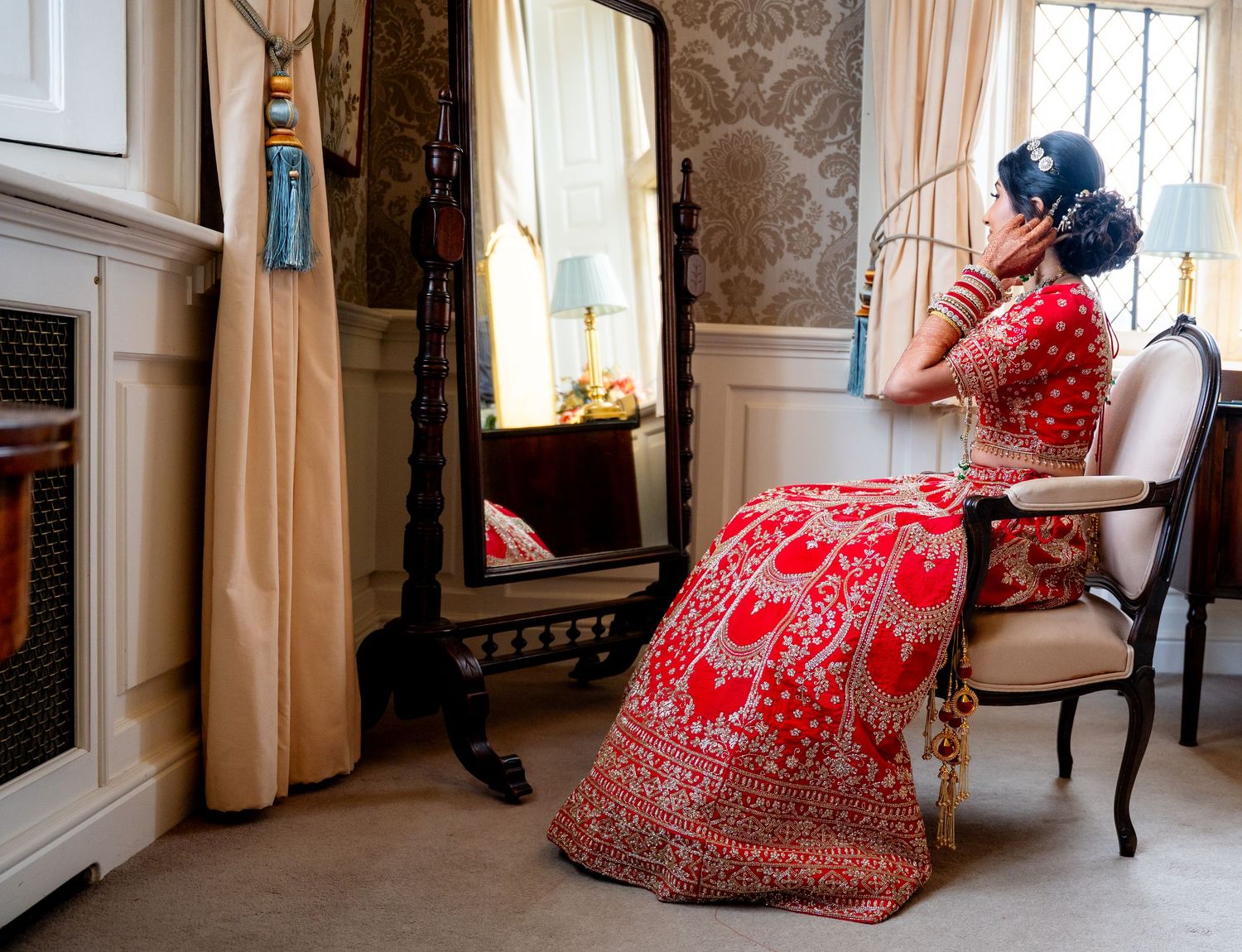 Indian bride in red lehenga sits before elegant mirror in luxurious bridal suite.
