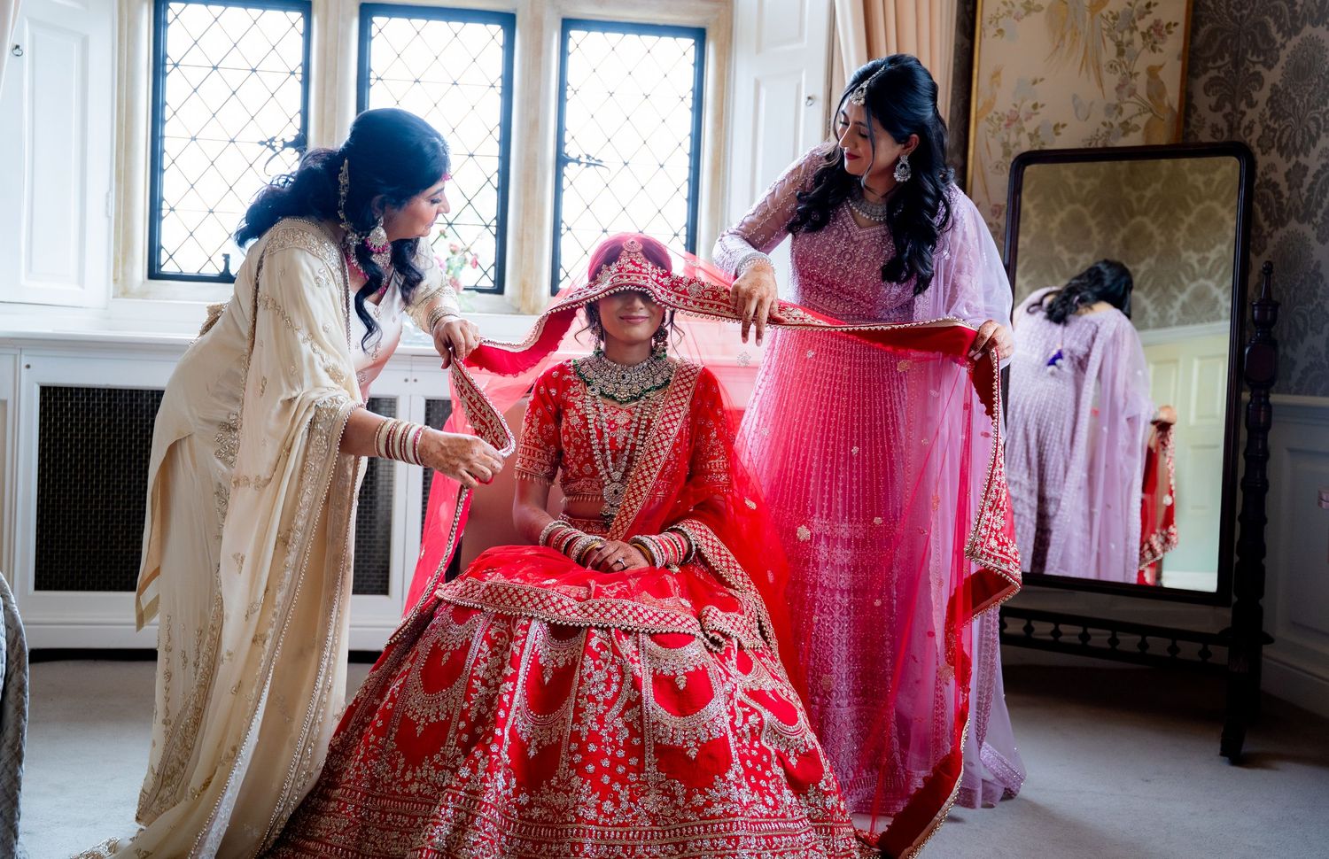 Bridesmaids in white and pink outfits helping Indian bride in red lehenga with her dupatta.