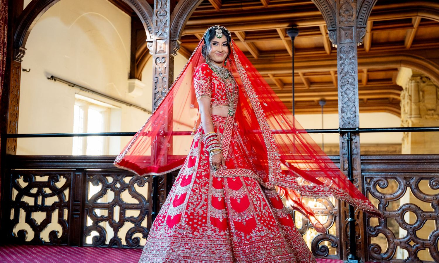 Stunning Indian bride in red embroidered lehenga posing in ornate venue.