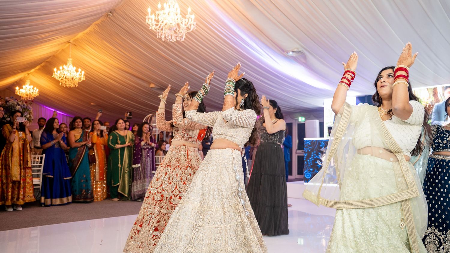 Women in colorful traditional Indian attire dance joyfully at an elegant wedding reception under chandelier lighting.