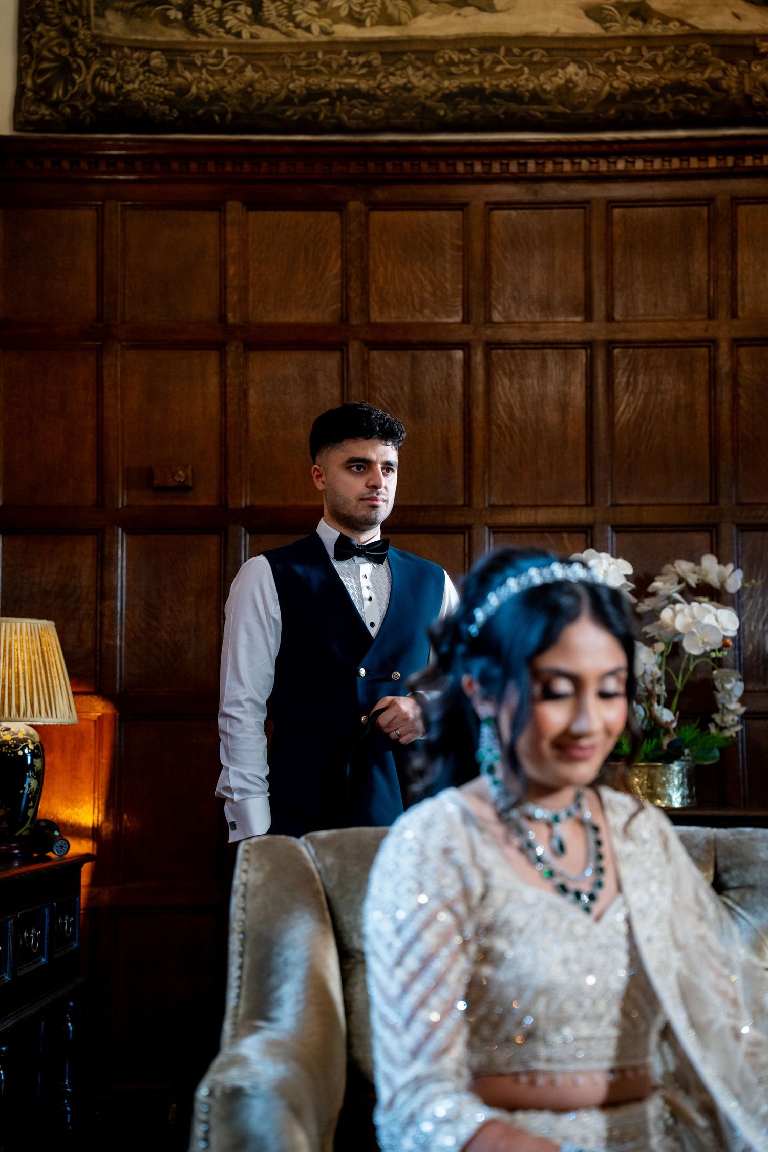 A groom in a navy waistcoat and bow tie stands behind his bride in a embellished white lehenga at an elegant wood-paneled venue.