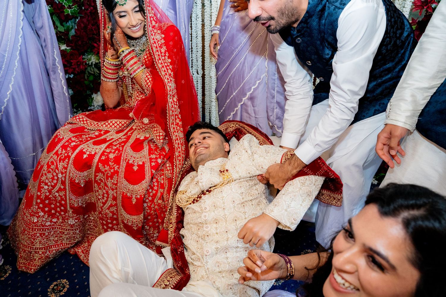 Groom playfully leans back during a joyful Indian wedding ceremony surrounded by laughing guests in traditional attire.